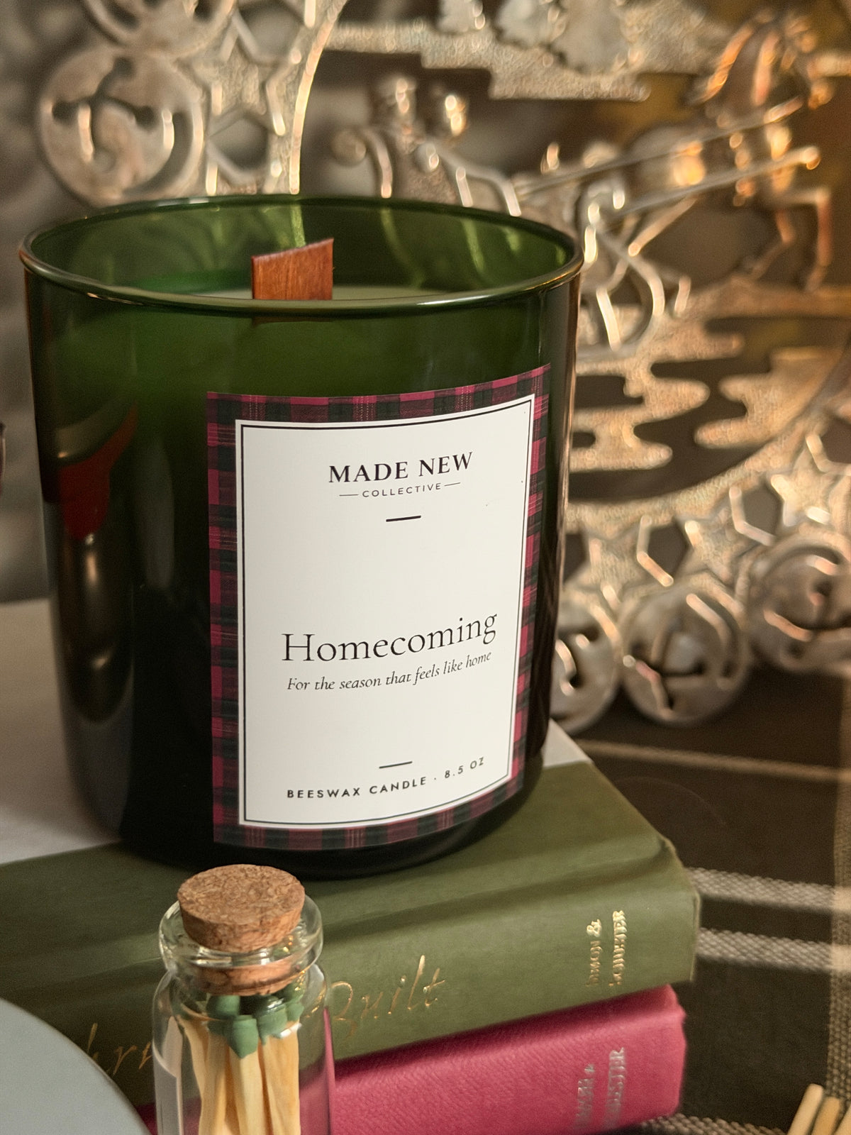 Close-up of the Homecoming candle label, plaid-bordered, on the green glass tumbler with wood wick, sitting on a vintage book next to a glass match jar