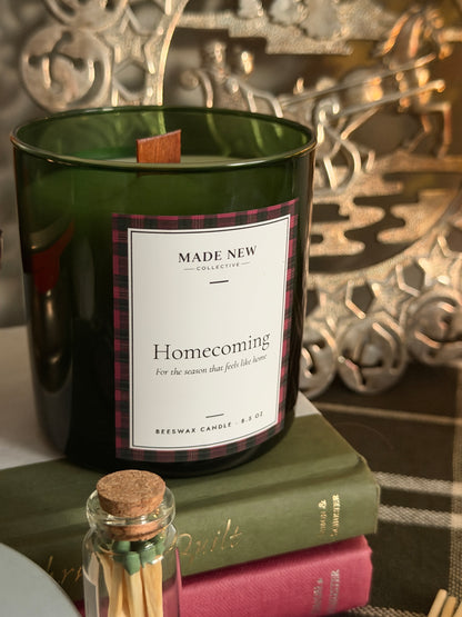 Close-up of the Homecoming candle label, plaid-bordered, on the green glass tumbler with wood wick, sitting on a vintage book next to a glass match jar