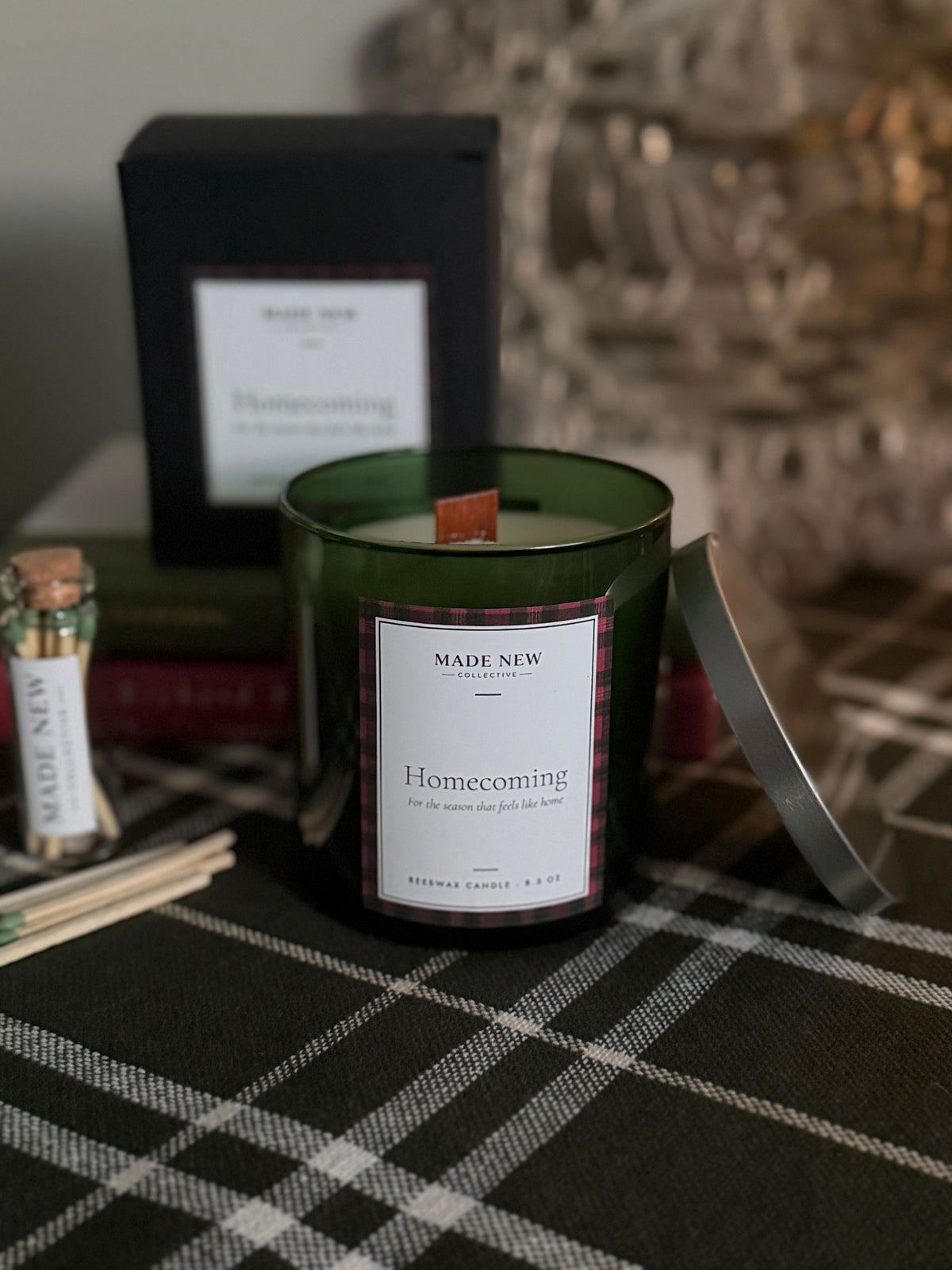 Homecoming holiday beeswax candle in a green glass tumbler with the silver lid off, black gift box and glass match jar beside it on a dark green plaid cloth — Made New Collective