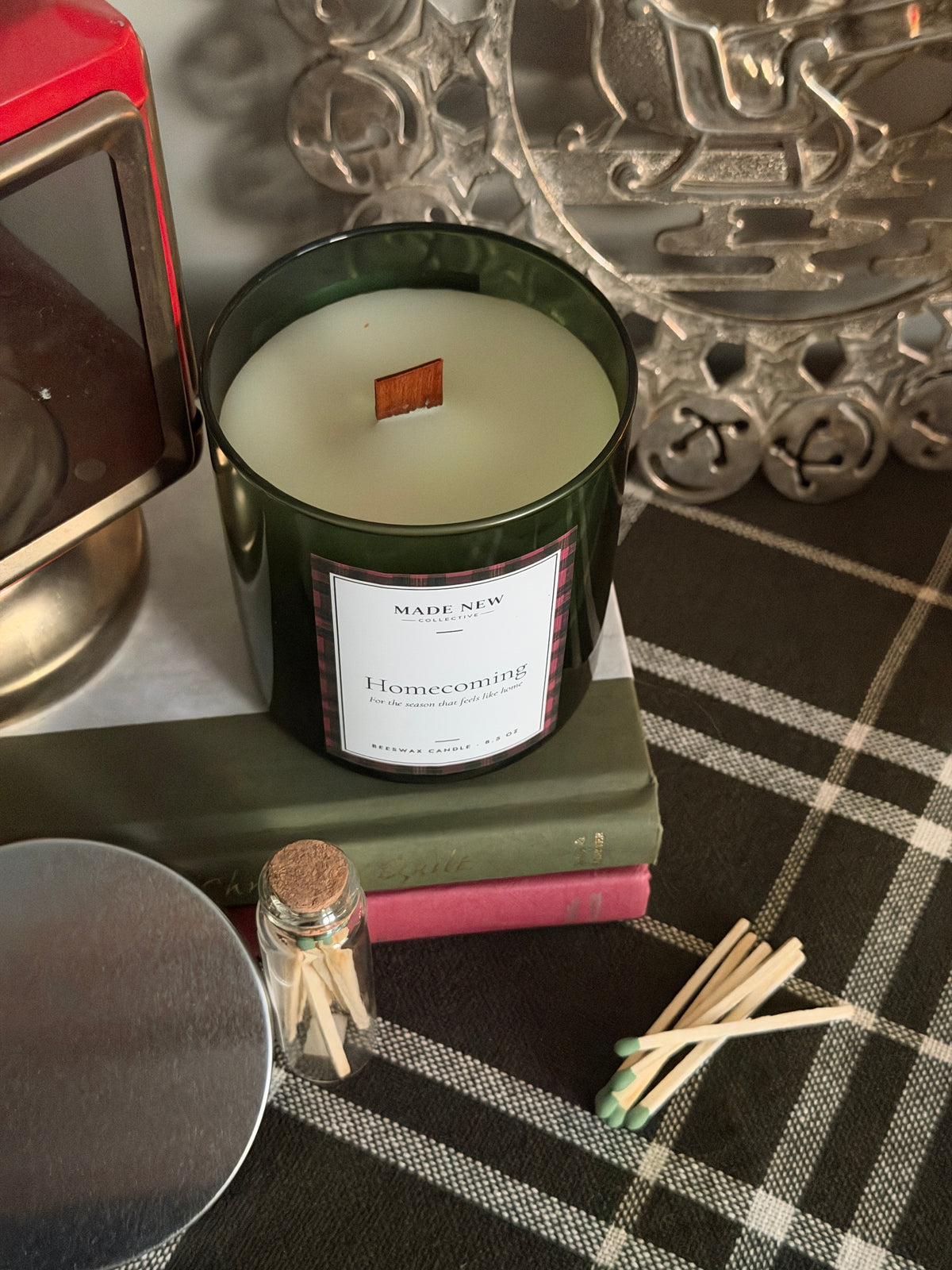 Homecoming candle seen from above with the silver lid set aside, resting on a vintage book on a green plaid cloth with loose matches and a Made New Collective match jar